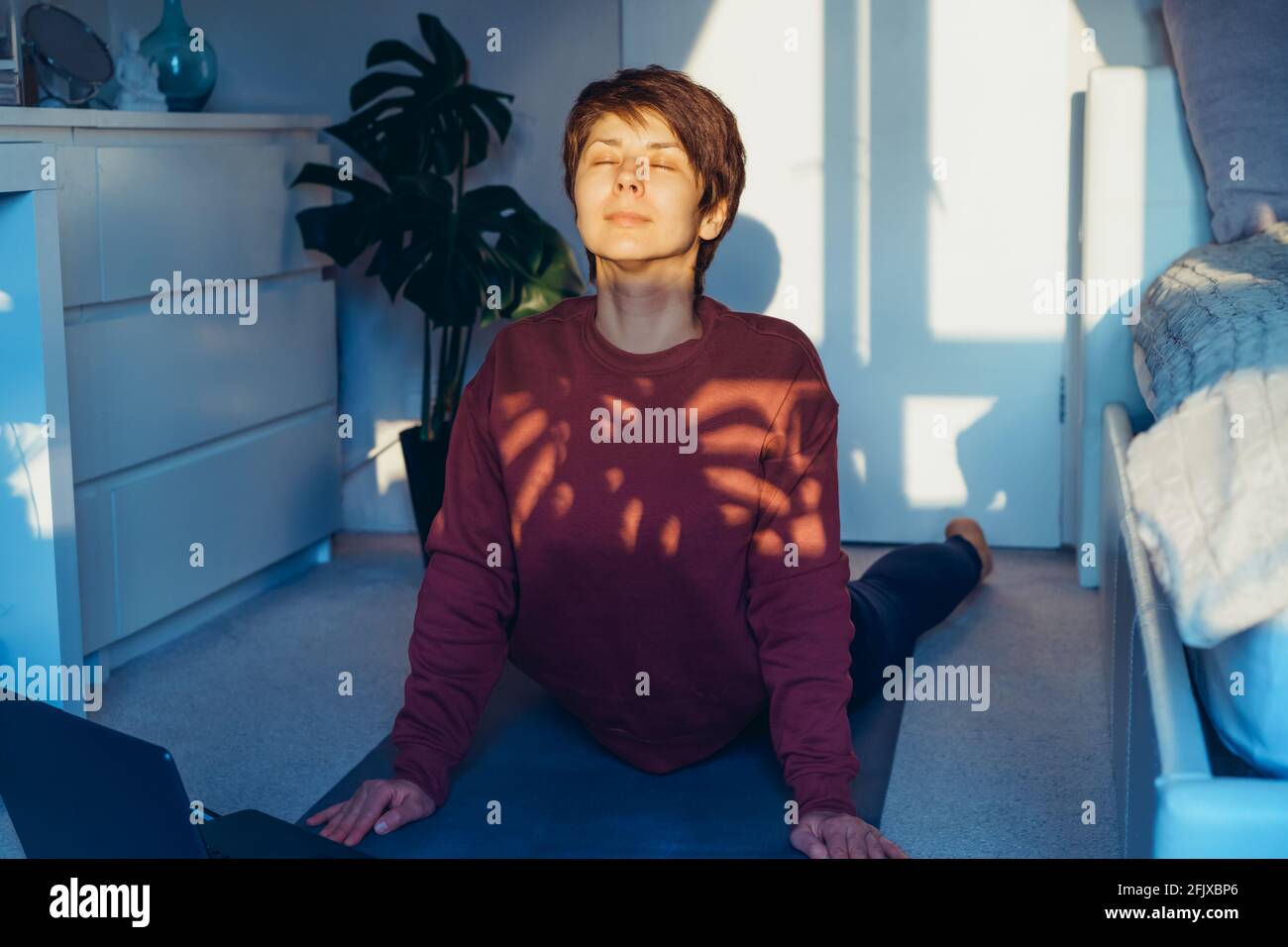 Frau mit gemischter Rasse und geschlossenen Augen, Yoga praktizierend, Hundeübung nach oben schauend, Urdhva Mukha svanasana Pose, mit dem sie in ihrem Schlafzimmer trainiere Stockfoto