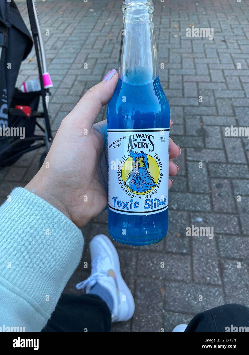 Eine blaue Soda-Pop-Flasche, die von einer Frau in weißen Nike Schuhen gehalten wird. Das altmodische Sodagetränk wird von der Marke Avery's Beverage als Toxic Slime bezeichnet Stockfoto