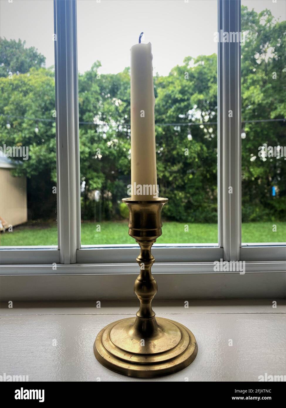 Weiße Kerze auf einem alten goldenen Messingleuchter, der durch ein Küchenfenster auf einen grünen Hof blickt. Stockfoto