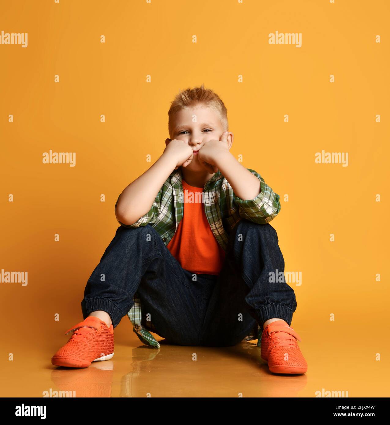 Gelangweilter Schüler Teenager, normaler Kerl im karierten Hemd, orangefarbenem T-Shirt und Jeans sitzt mit Fäusten an seinen Wangen Stockfoto