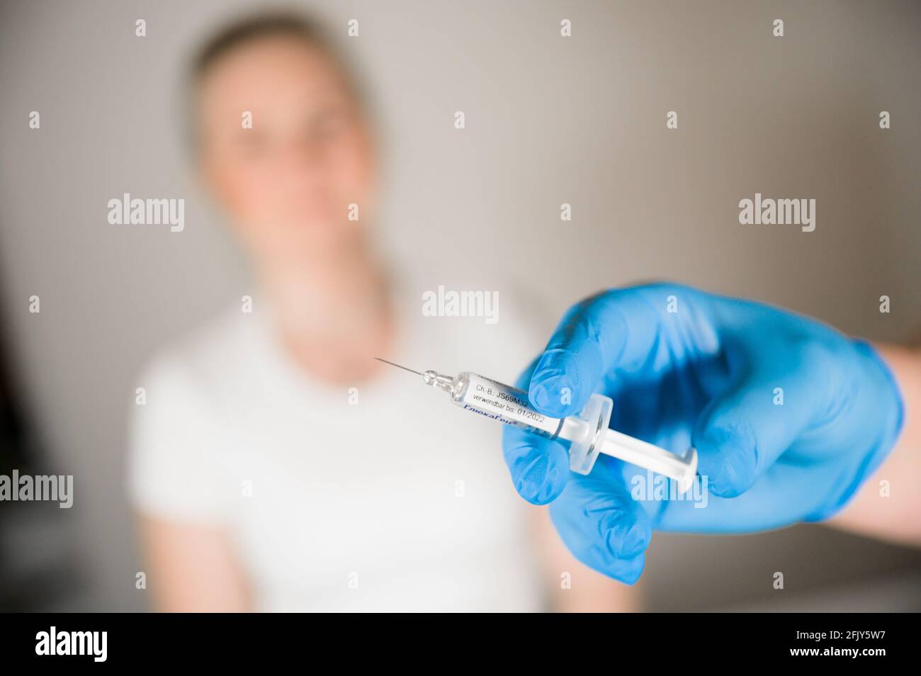 Arzt, der einen Covid-Impfstoff in den Händen hält, mit einer jungen blonden Patientin im Hintergrund: Heilung der Corona-Pandemie durch Massenimpfung Stockfoto