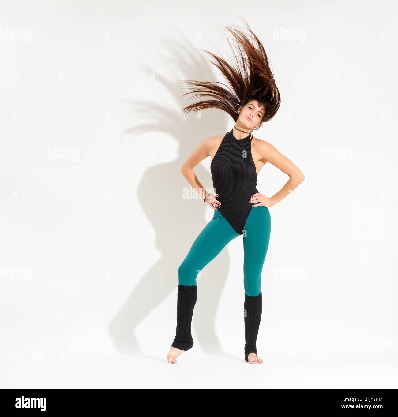 Formschöne junge Tänzerin in einem 80er-Jahre-Outfit, die einen Klassiker aufführt Retro-Tanzpose mit Händen auf Hüften, die sie lang werfen Haare hoch in die Luft isoliert auf w Stockfoto