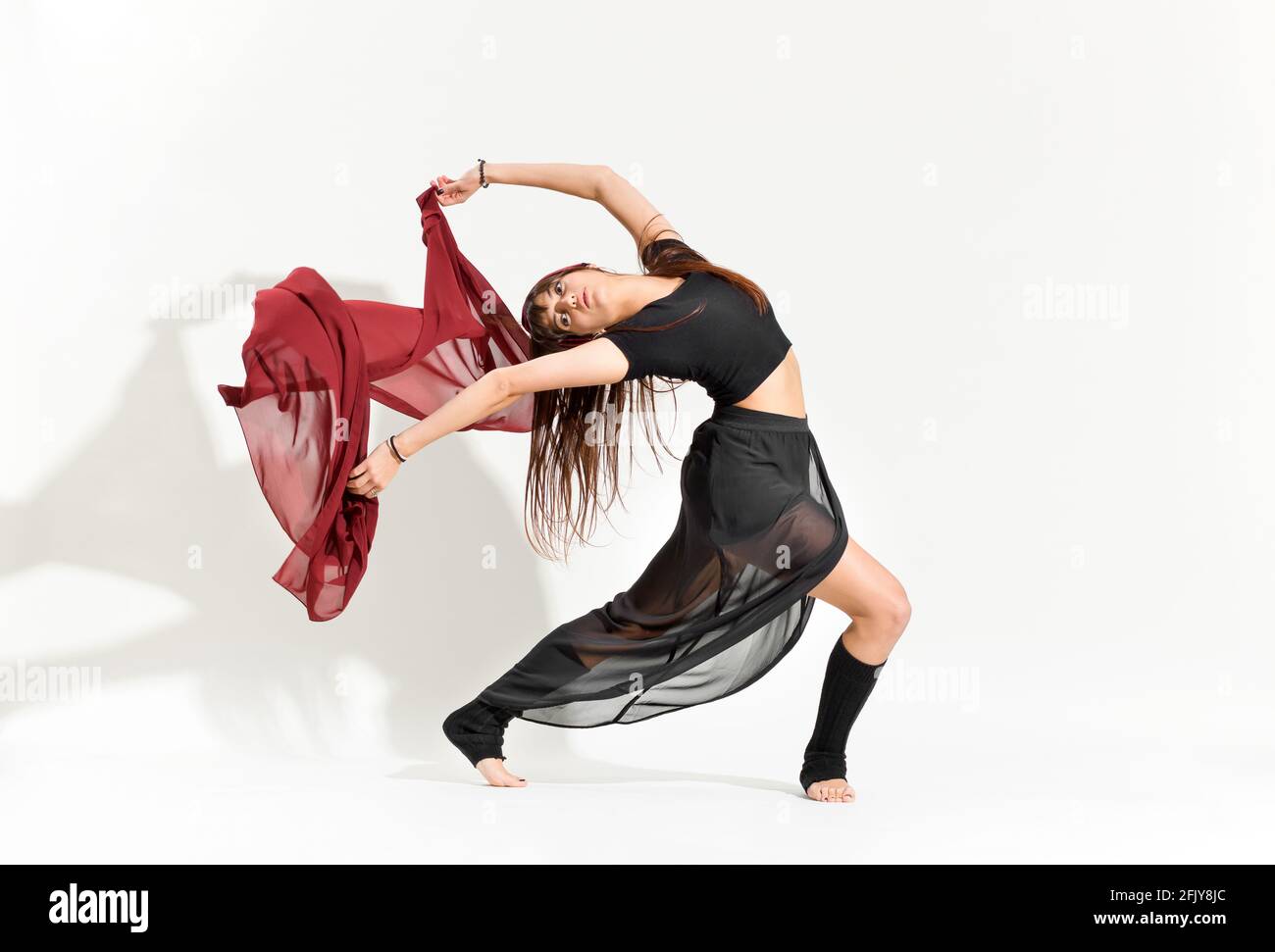 Anmutiger Tanz, der eine Ina Bauer-Eiskunstlauf-Pose vorführt Ein filmiges schwarzes Kostüm, das ein Rechteck aus rotem Stoff schwebt Hinter ihr auf weiß mit Schatten Stockfoto
