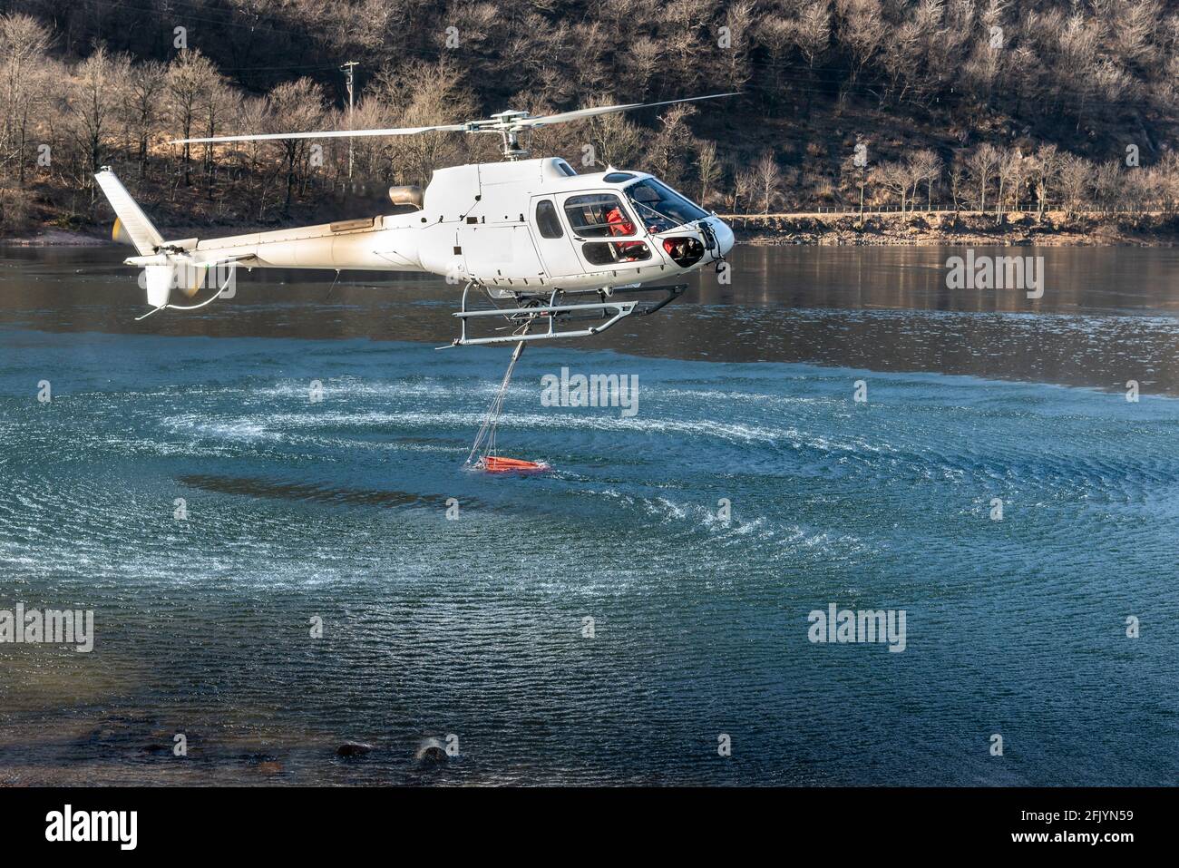 Der Hubschrauber lädt Wasser aus dem Ghirla-See, um die Bergflammen in Valganna, Italien, zu löschen. Der Kampf gegen Waldbrände. Stockfoto