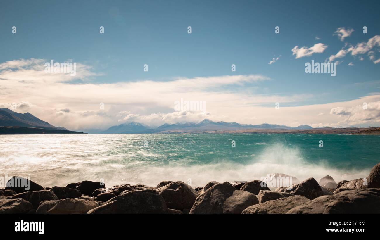 Starker Wind und krachende Wellen treffen auf die Felsen am See Pukaki mit Mt Cook bedeckt von den Wolken im Entfernung Stockfoto