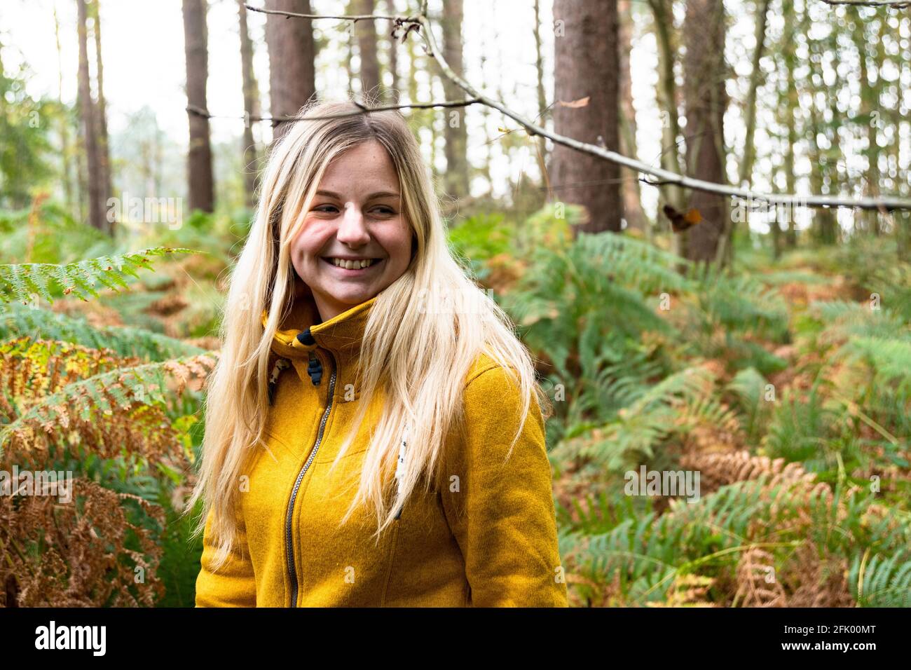 Blonde Frau in gelber Jacke, die durch eine Waldlichtung geht Mit Farn überwuchert Stockfoto