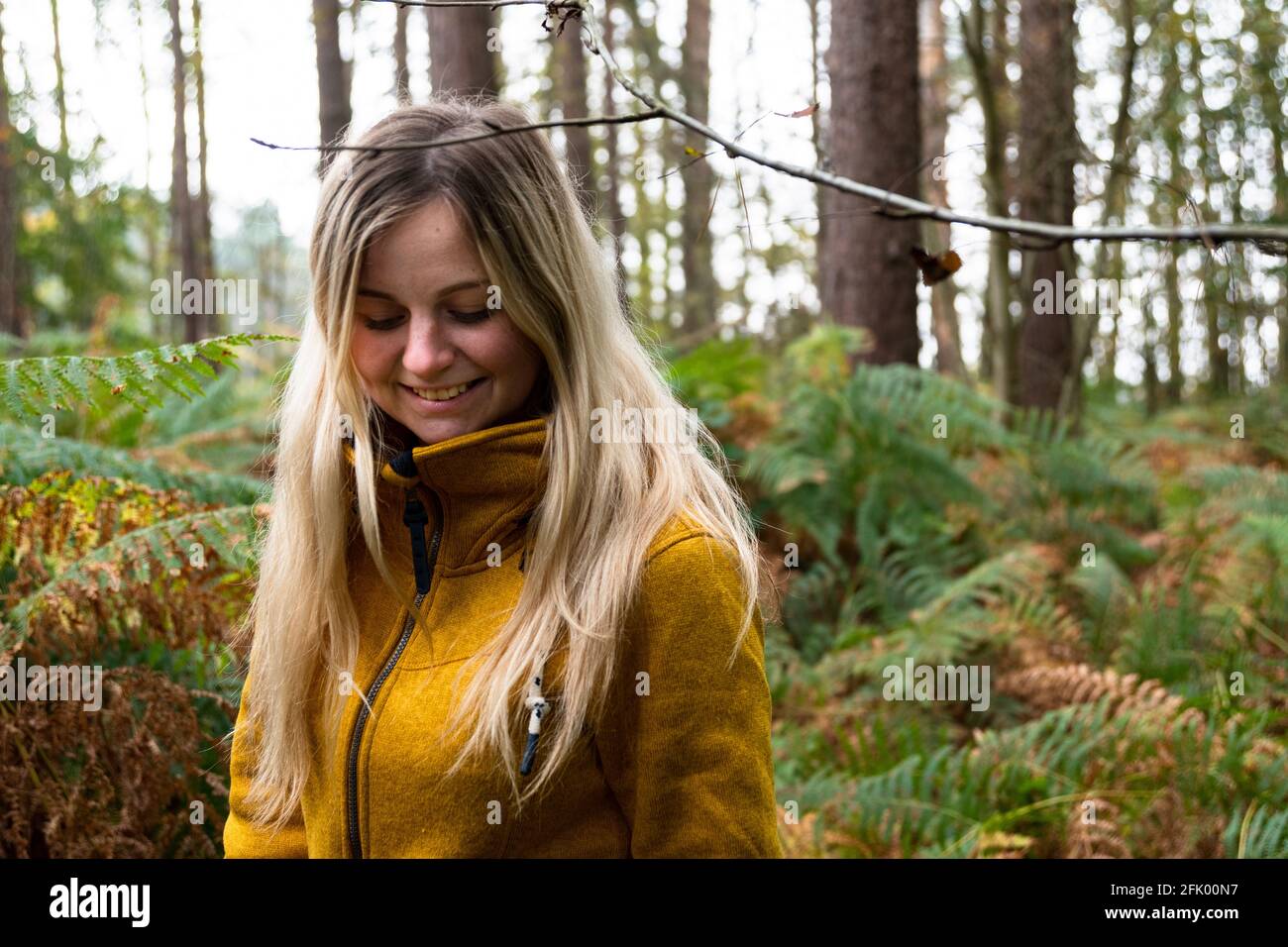 Blonde Frau in gelber Jacke, die durch eine Waldlichtung geht Mit Farn überwuchert Stockfoto