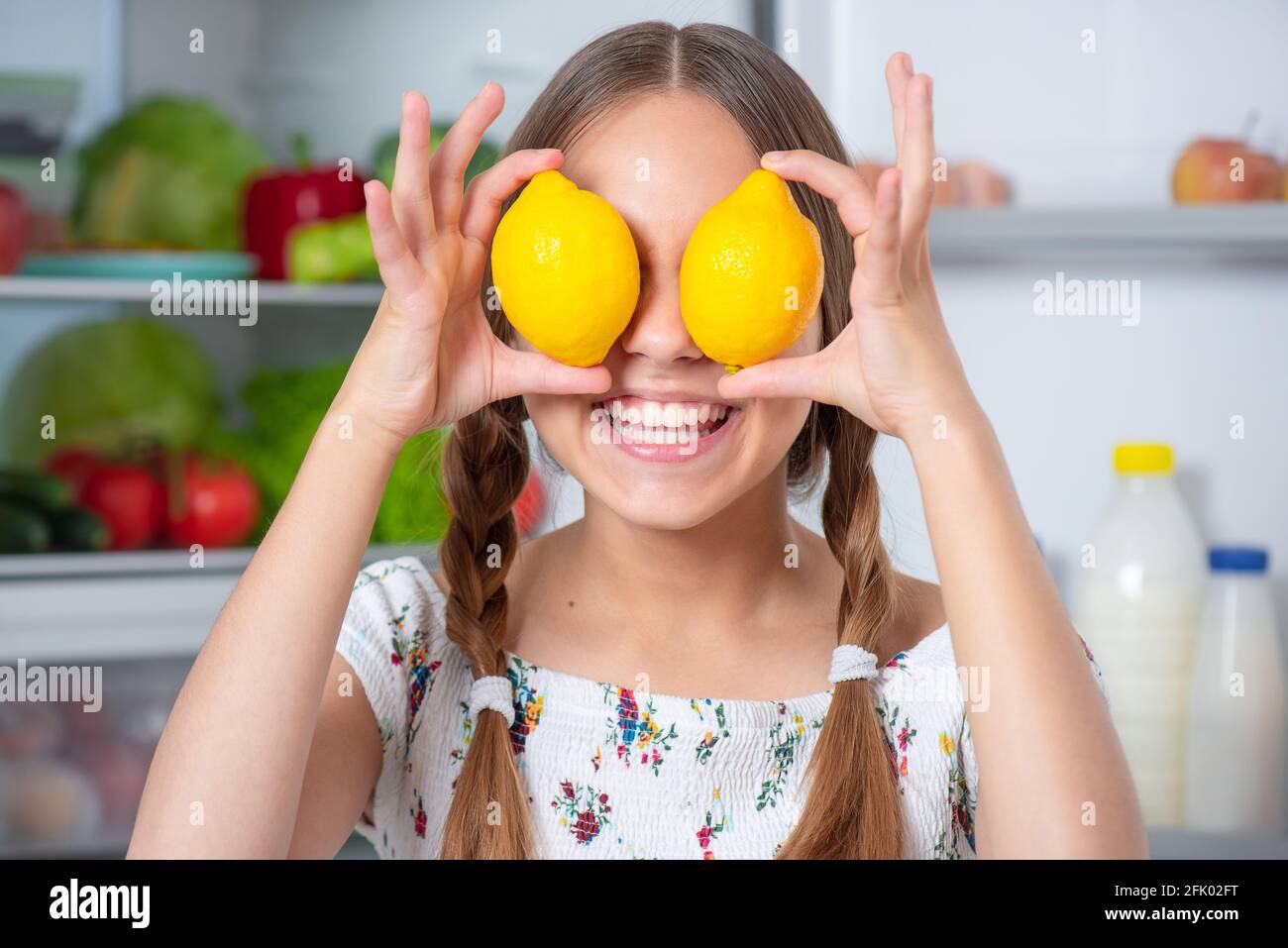 Lächelnd schöne junge Teenager-Mädchen hält gelbe Zitronen über den Augen Während Sie in der Nähe des offenen Kühlschranks in der Küche zu Hause stehen Stockfoto