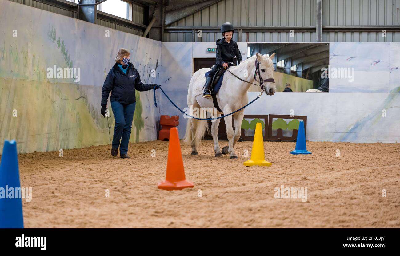 East Lothian, Schottland, Großbritannien, 27. April 2021. Muirfield Riding Therapy wird wieder eröffnet: Die Wohltätigkeitsorganisation begrüßt ihre ersten Jugendlichen zu ihrer Therapiestunde mit Lockdown-Einschränkungen. Aufgrund der anhaltenden Einschränkungen können derzeit nur die mobilen Clients bedient werden, die immer mehr Kunden betreuen. Im Bild: Nathan, 10 Jahre alt, ein Schüler an der Yester Primary School auf seinem Therapiepferd Johnny unter der Leitung der Freiwilligen Susan, der lernt, die Zügel zu kontrollieren und das Pferd zu lenken Stockfoto