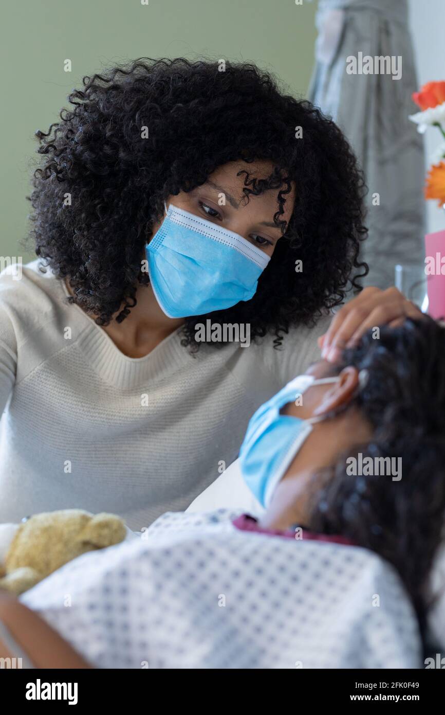 Mutter mit gemischter Rasse und kranke Tochter in Gesichtsmasken im Krankenhaus, Mädchen mit Teddybär Stockfoto