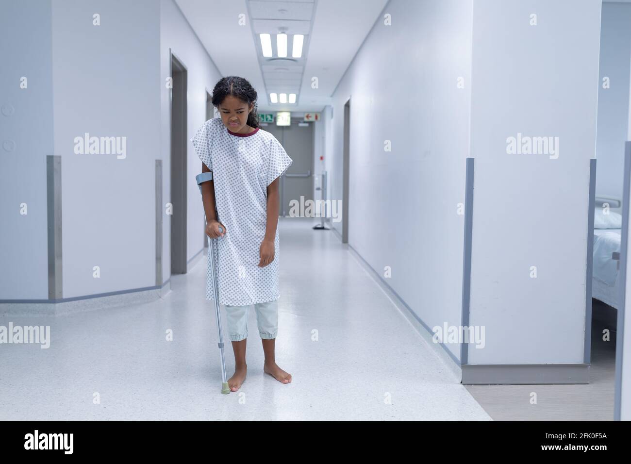 Krank gemischtes Rennmädchen, das barfuß im Krankenhauskorridor läuft Eine Krücke Stockfoto