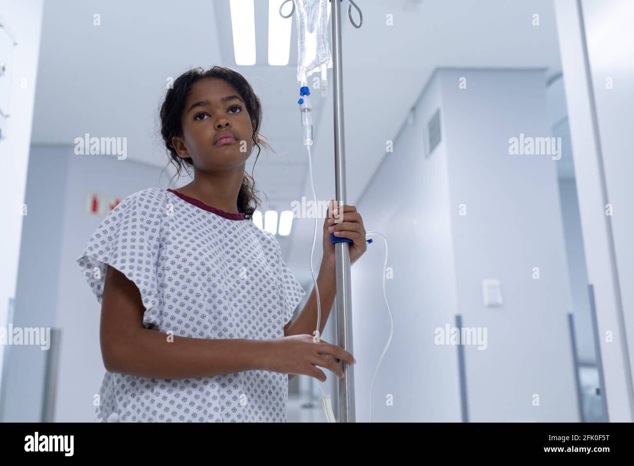 Krankes Mädchen mit gemischter Rasse, das im Krankenhausflur mit iv läuft Tropfbeutel auf Ständer Stockfoto