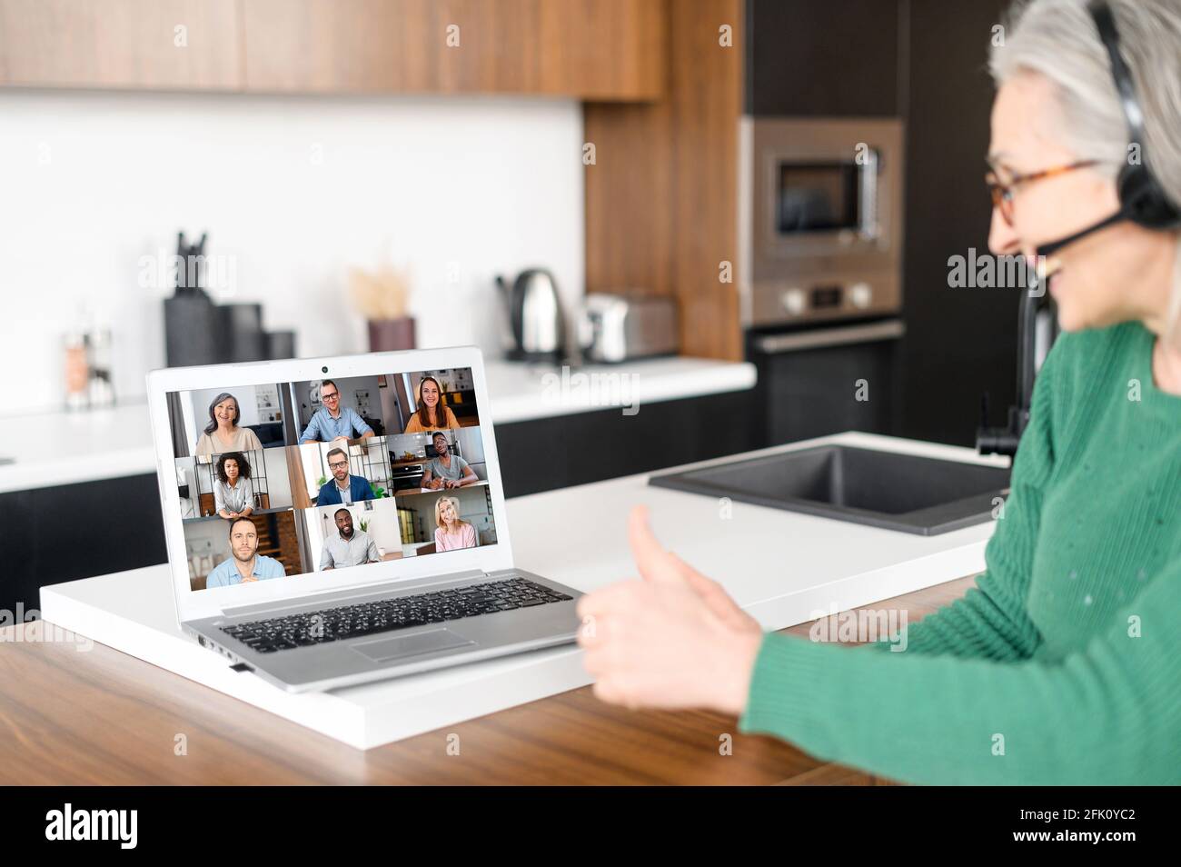 Rückansicht über die Schulter einer älteren Frau auf dem Laptop-Bildschirm mit vielen verschiedenen Personen, die auf dem Laptop leben, hat ein reifer Mitarbeiter, der ein schnurloses Headset für virtuelle Meetings mit Kollegen verwendet, ein virtuelles Meeting Stockfoto
