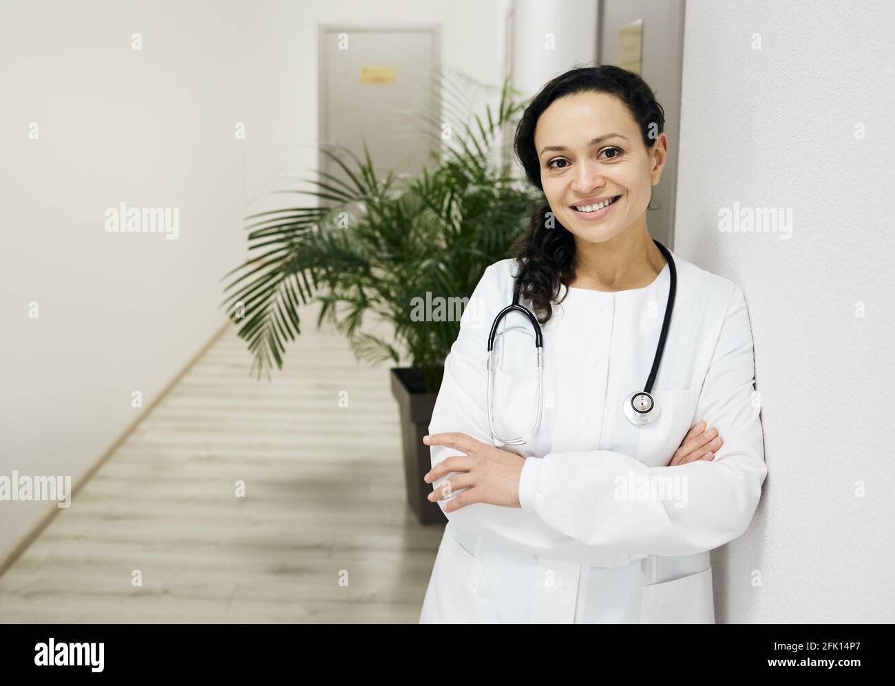 Allgemeinmediziner. Positive Ärztin mit gemischter Rasse, die eine medizinische Uniform mit gekreuzten Armen auf dem Krankenhausflur trägt Stockfoto