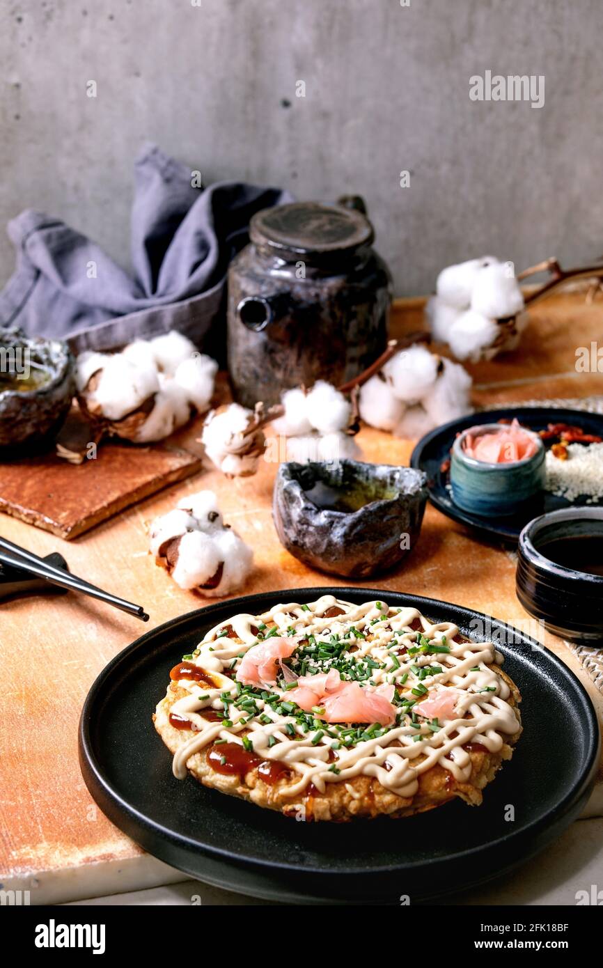 Hausgemachte japanische Fast Food Okonomiyaki Kohl Pfannkuchen mit Zwiebel, eingelegter Ingwer, Mayo-Sauce auf schwarzem Keramikplatte. Essstäbchen, Teekanne, Baumwolle, ING Stockfoto