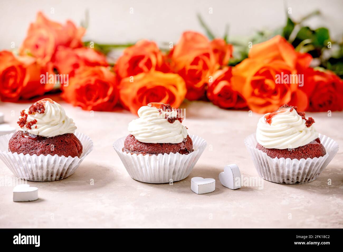 Hausgemachte Cupcakes aus rotem Samt mit Schlagsahne in Reihe, weiße Serviette mit Band, Rosenblumen, Holzherzen auf rosa Textur-Hintergrund. Valentinstag Stockfoto