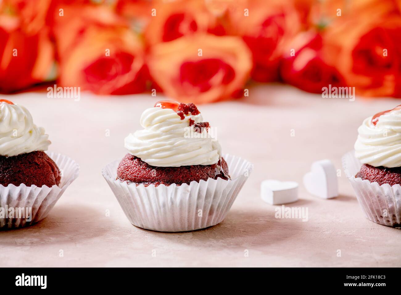 Hausgemachte Cupcakes aus rotem Samt mit Schlagsahne in Reihe, weiße Serviette mit Band, Rosenblumen, Holzherzen auf rosa Textur-Hintergrund. Valentinstag Stockfoto