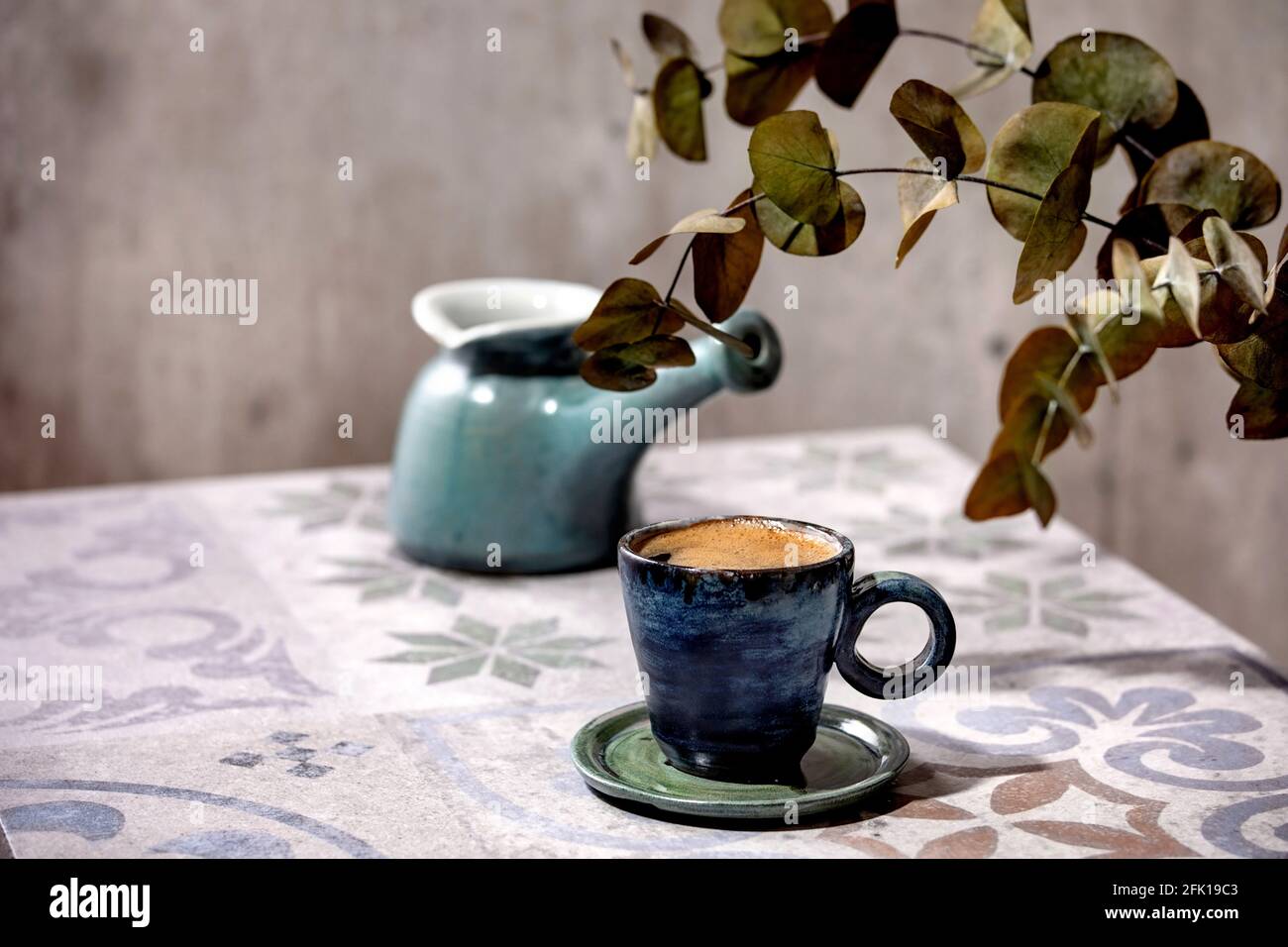 Tasse türkischer schwarzer Schaumkaffee auf einem kunstvollen Keramiktisch mit Kaffeekanne, Cezve und Eukalyptuszweigen. Sonnenlicht Stockfoto