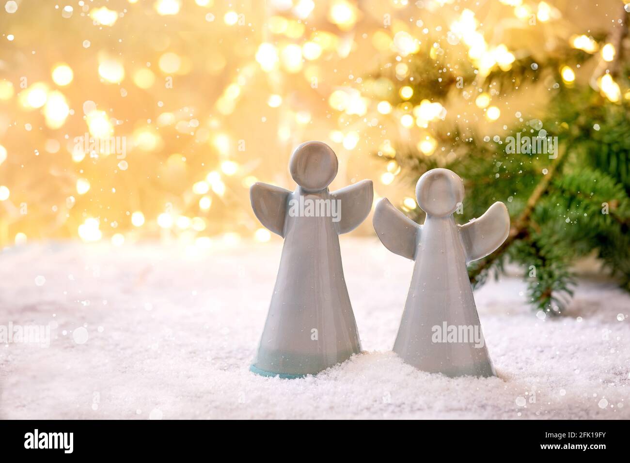 Weihnachtsengel aus Keramik. Set aus zwei handgefertigten Weihnachtsschmuck Engel auf Schnee mit Bokeh Weihnachtsbeleuchtung und Tannenzweigen. Stockfoto