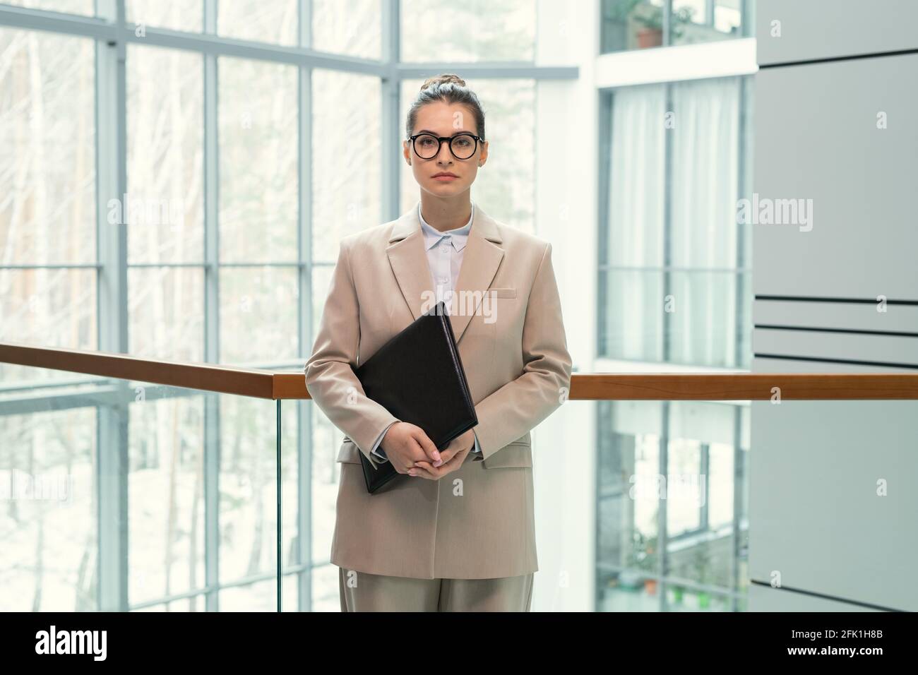 Porträt einer seriös attraktiven Geschäftsfrau in runden Brillen stehend mit Schwarzer Ordner in der Lobby des Bürozentrums Stockfoto