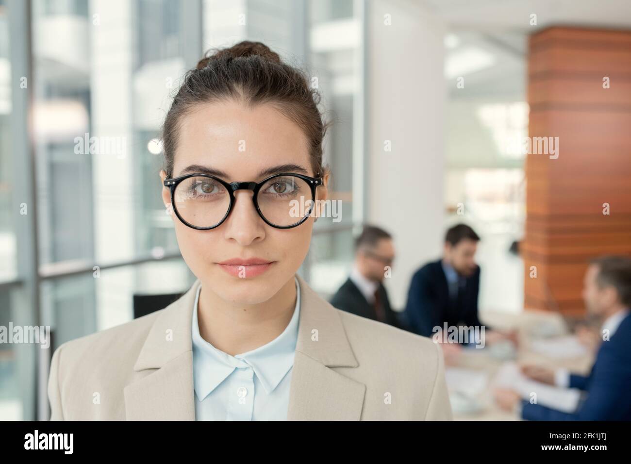 Porträt einer selbstbewussten attraktiven Rechtsanwältin in runden Brillen stehend Gegen Kollegen, die im Hintergrund über Papiere diskutieren Stockfoto