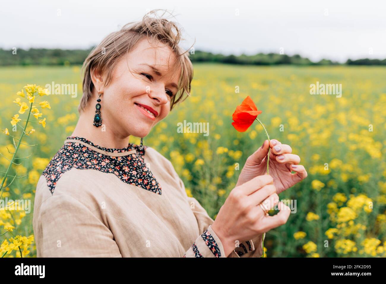 Horizontales Porträt einer attraktiven jungen Frau auf einem Blumenfeld. Sie lächelt, während sie eine Mohnblume hält Stockfoto