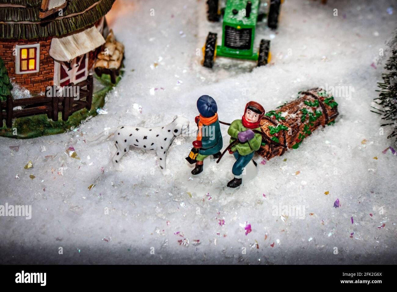 Yule Log wird von zwei Jungen im Weihnachtsdorf geschleppt Szene durch tiefen Schnee mit Traktor und Windmühle verschwommen hinter Und Dalmatiner Hund schnüffelt bo Stockfoto