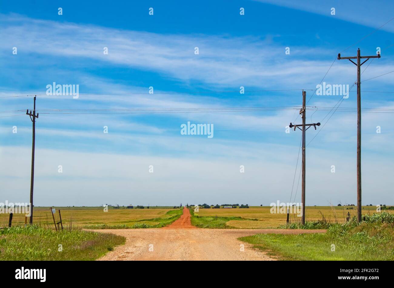 Die Straße weniger gereist - eine ländliche Kreuzung mit Eine rote Schotterstraße, die nach vorne über den Horizont führt Farmland unter sehr blauem Himmel Stockfoto