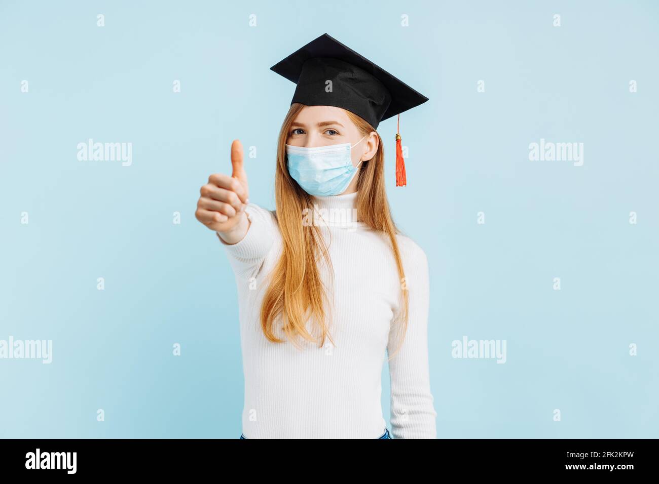 Glücklich weibliche Student Absolvent in medizinische Maske zeigt Daumen nach oben Auf blauem Hintergrund Stockfoto