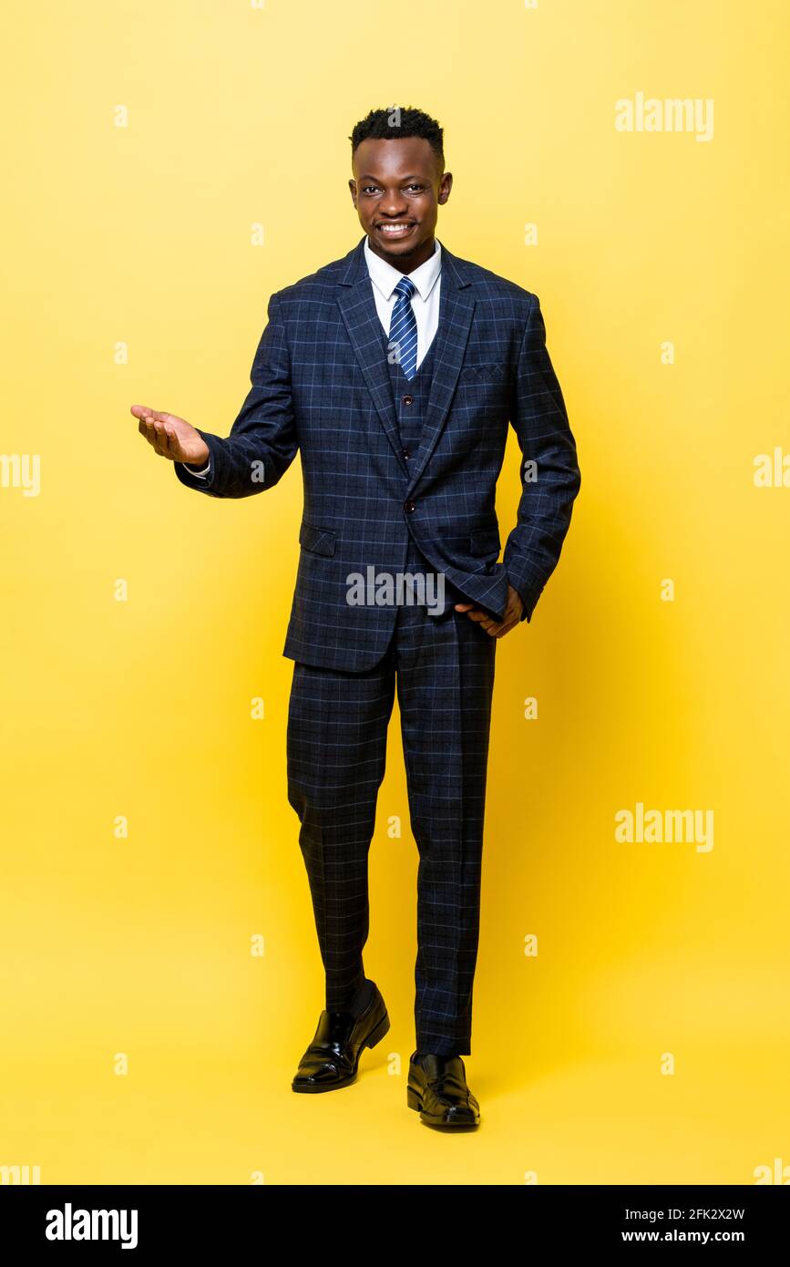 In voller Länge Porträt eines jungen hübschen afrikanischen Mannes im Geschäft Anzug lächelt und öffnet seine Handfläche in einem gelben, isolierten Studio Hintergrund Stockfoto