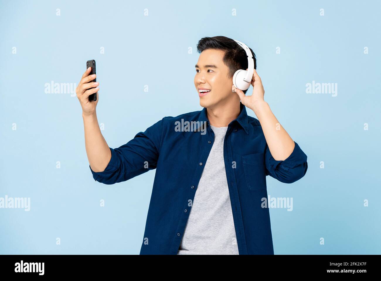 Glücklich lächelnder asiatischer Mann trägt Kopfhörer und hört Musik von Mobiltelefon im hellblauen, isolierten Studiohintergrund Stockfoto