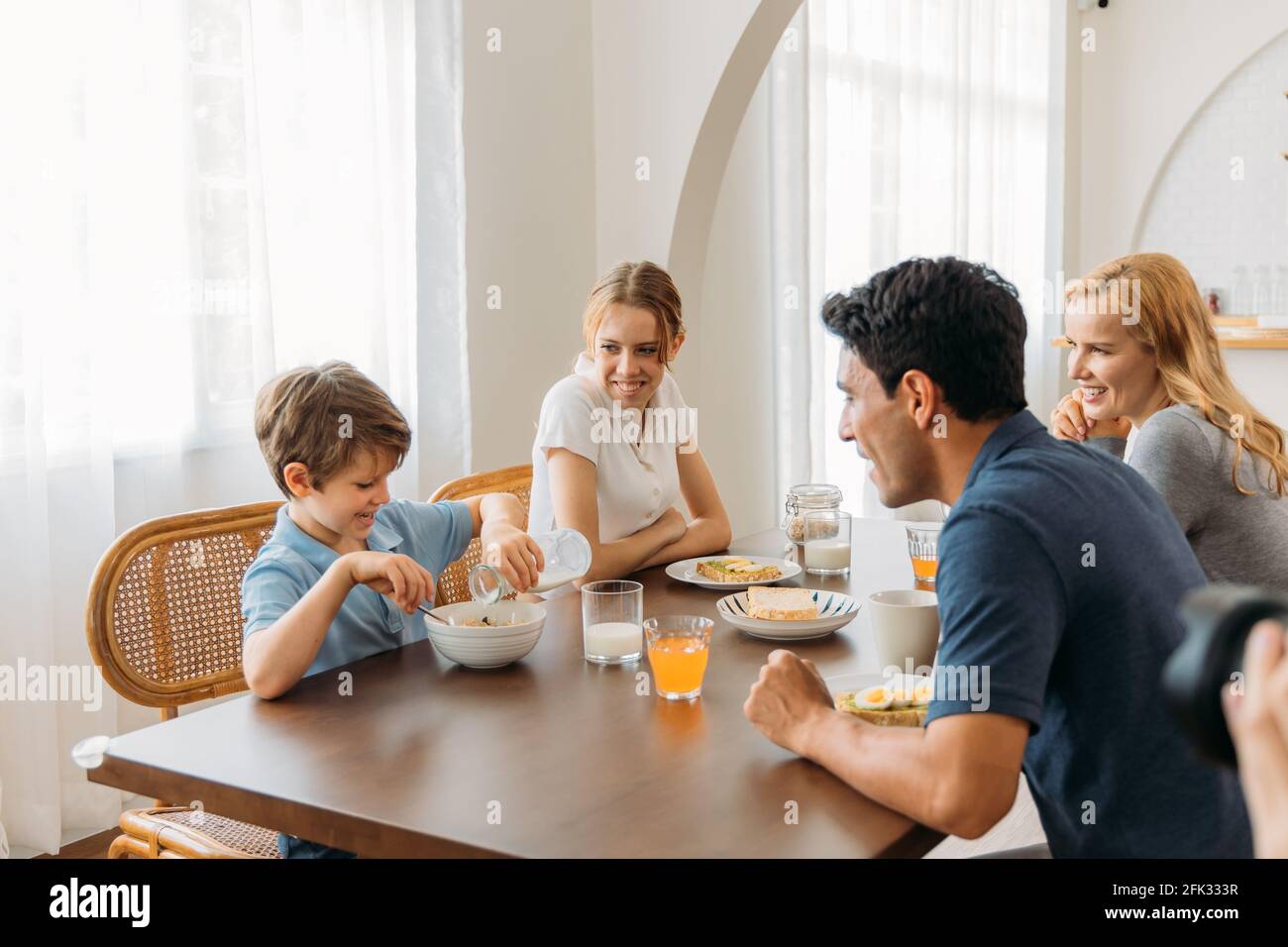 Schöner junger Vater zusammen mit schöner Mutter und Teenager-Tochter Ich schaue auf einen kleinen Sohn, der Milch aus der Flasche in die Schüssel gießt Von Cerealien während des Frühstücks zu Hause Stockfoto