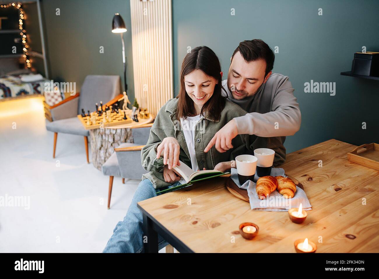 Schönes Paar mittleren Alters, das neben einem Frühstückstisch ein Buch mit Kaffee und Croissants las. Sie sitzen auf einem Stuhl. Er zeigt auf einige s. Stockfoto