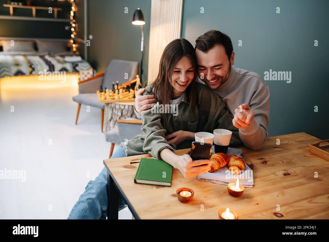 Ein fröhliches Paar mittleren Alters, das ein Selfie neben einem Frühstückstisch mit Kaffee und Croissants gemacht hat. Sie sitzen auf einem Stuhl. Kerzen werden angezündet Stockfoto