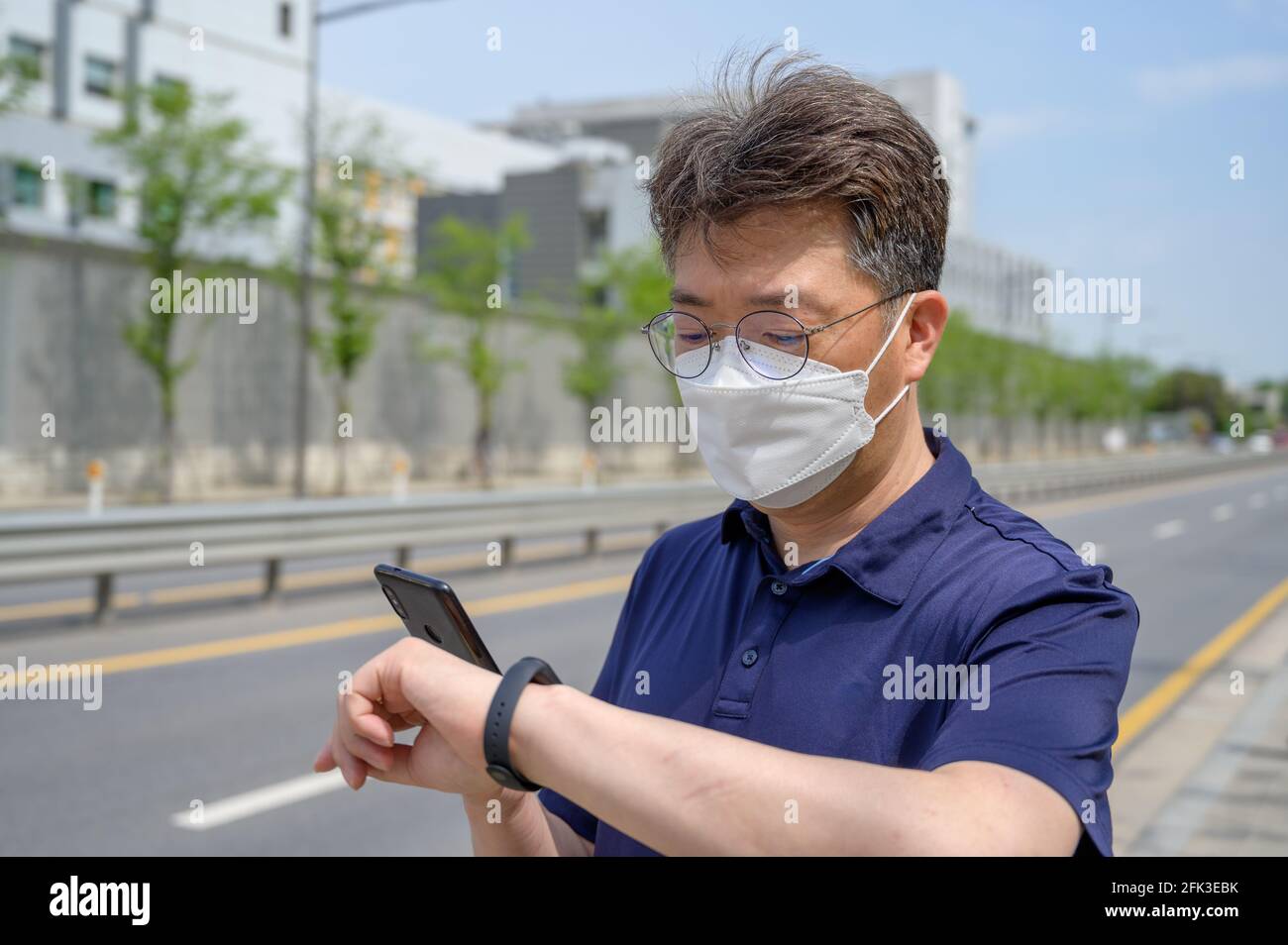 Asiatischer Mann mittleren Alters, der eine Gesichtsmaske trägt und ein Smartphone in der Auffahrt anschaut. Stockfoto