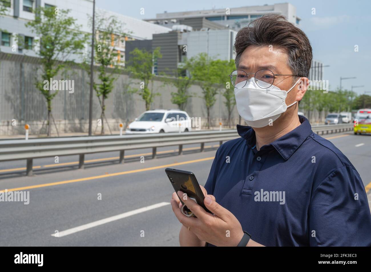 Asiatischer Mann mittleren Alters, der eine Gesichtsmaske trägt und ein Smartphone in der Auffahrt anschaut. Stockfoto