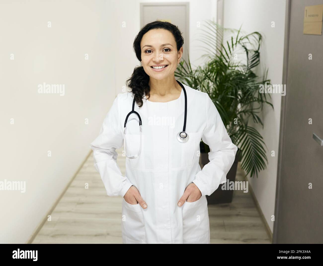 Allgemeinmediziner. In einem Krankenhausflur steht eine lächelnde Ärztin mit gemischter Rasse, die eine medizinische Uniform mit einem Stethoskop trägt Stockfoto