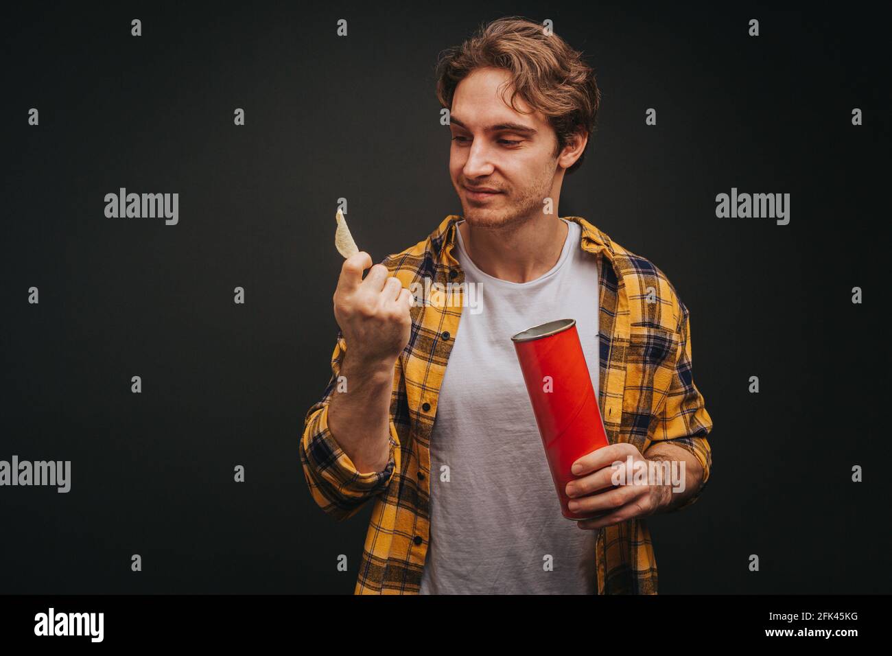 Der junge blonde Mann in gelbem Hemd hält Chips, isoliert auf schwarzem Hintergrund Stockfoto
