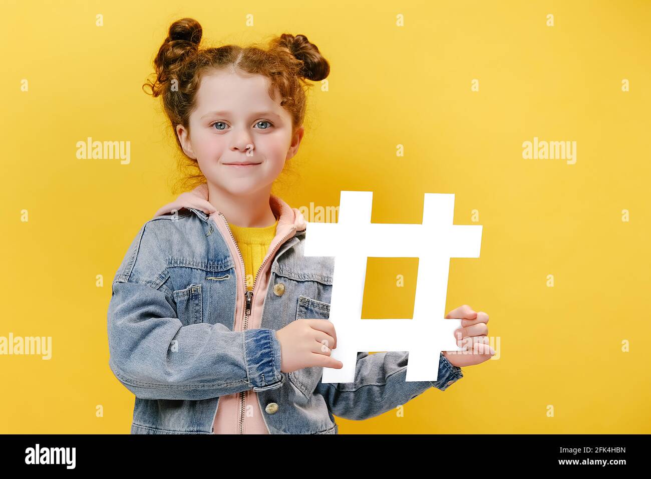 Süßes Mädchen Kind hält großen weißen Hashtag und glücklich Blick auf die Kamera, trägt Denim-Jacke, isoliert über gelben Studio-Hintergrund mit Kopierraum Stockfoto