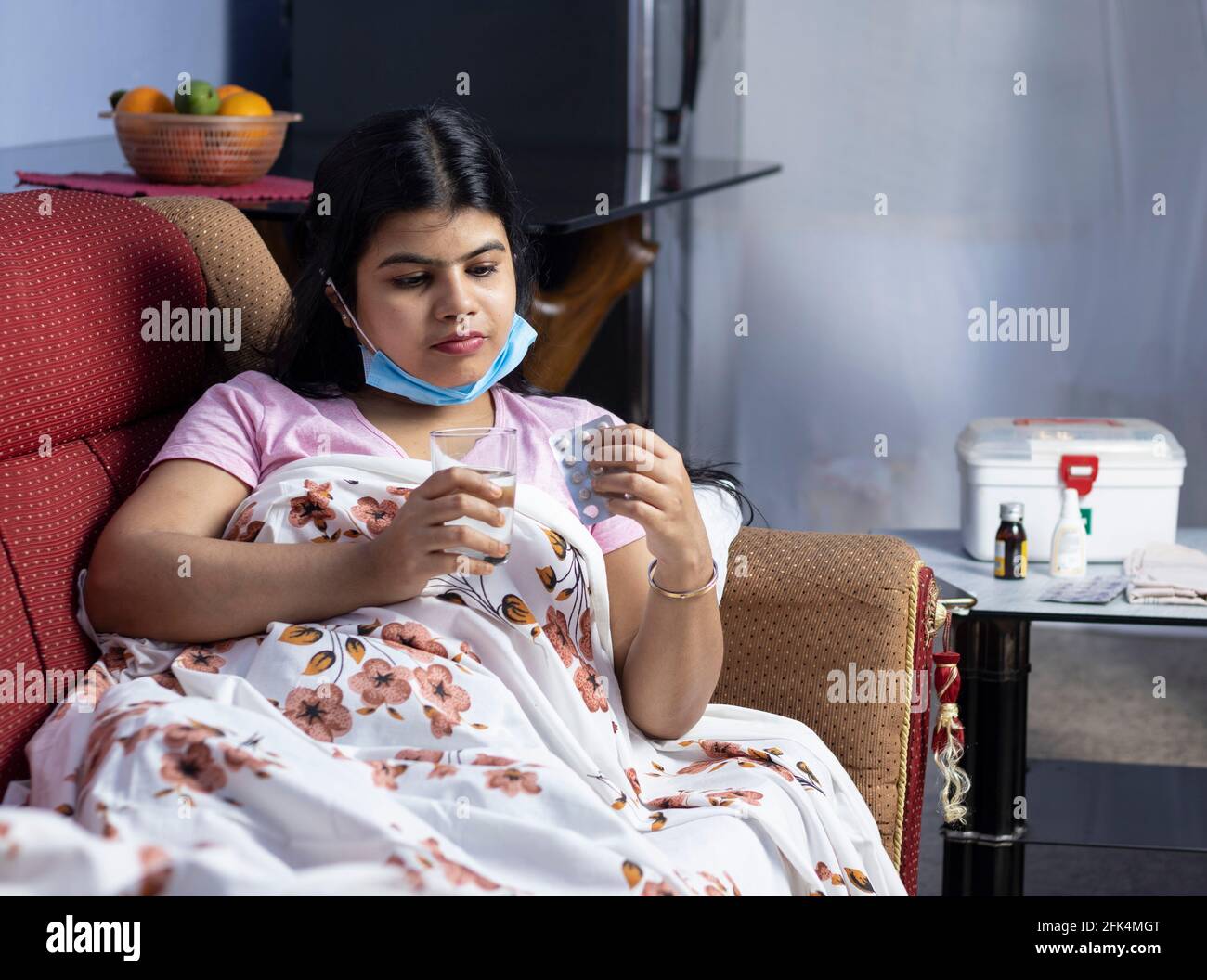 Eine indische Frau mit mittlerem Erwachsenen, die eine chirurgische Nasenmaske trägt, auf dem Sofa liegt, Medikamente nimmt, Gesundheitskonzept Stockfoto