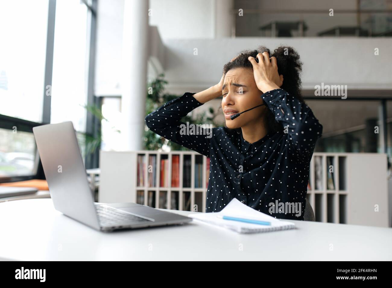 Verärgert müde afroamerikanische junge lockige Frau, mit Headset, Manager, Callcenter-Mitarbeiter oder Support-Operator, sitzt am Schreibtisch, fühlt sich gestresst, hält den Kopf mit den Händen und schreit Stockfoto