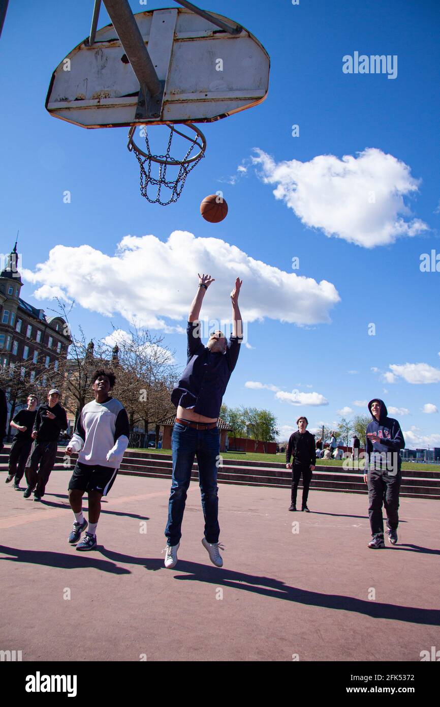 Multiethnische junge Menschen zahlen Straßenkbasketball. Der Ball liegt in der Luft und die Spieler werden ausgespielt. Blauer Himmel und Gebäude im Hintergrund. Kopenhagen Stockfoto