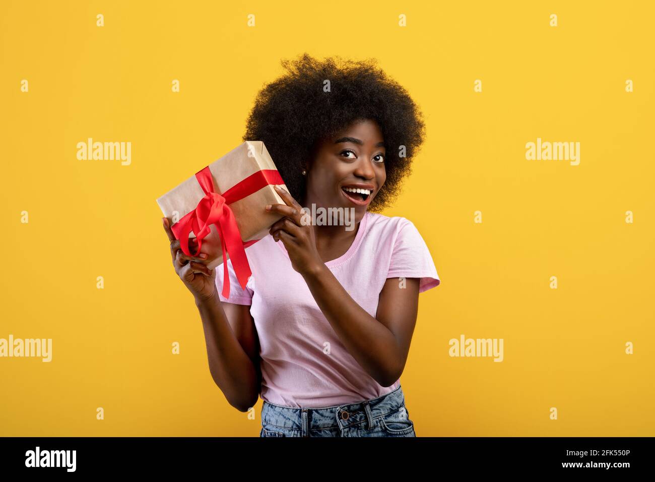 Erraten Sie, was drin ist. Glückliche schwarze Frau hört, was klappert in Geschenkbox, gelben Studio-Hintergrund Stockfoto