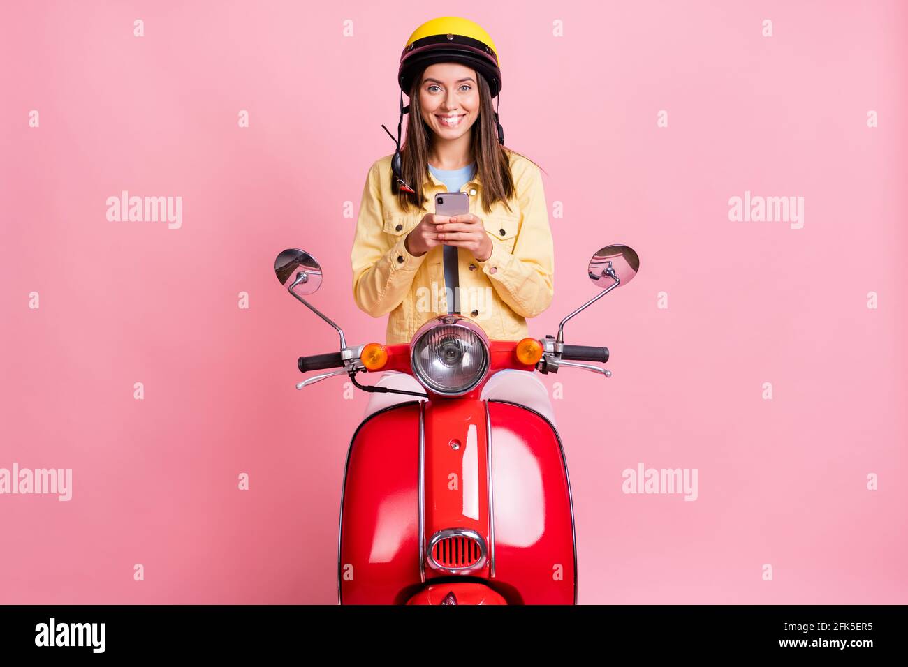 Foto von jungen lächelnd positive Mädchen Frau chatten Telefon tragen Gelber Helm Fahrt rotes Motorrad isoliert auf rosa Hintergrund Stockfoto