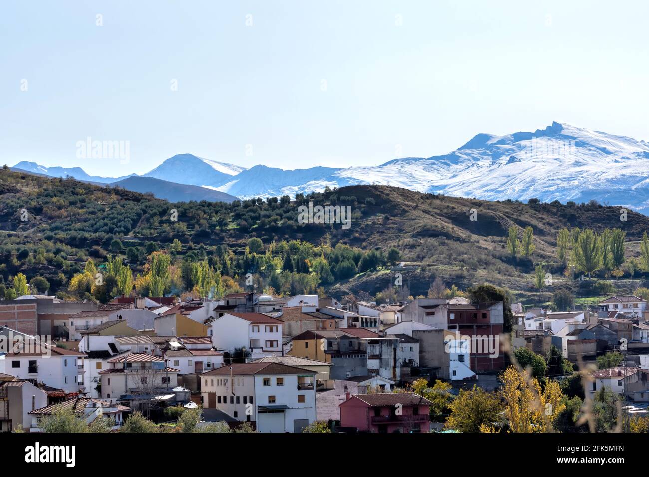 Blick auf die andalusische Stadt Beas de Granada inmitten von Bäumen Mit den hohen schneebedeckten Gipfeln der Sierra Nevada in der Hintergrund Stockfoto