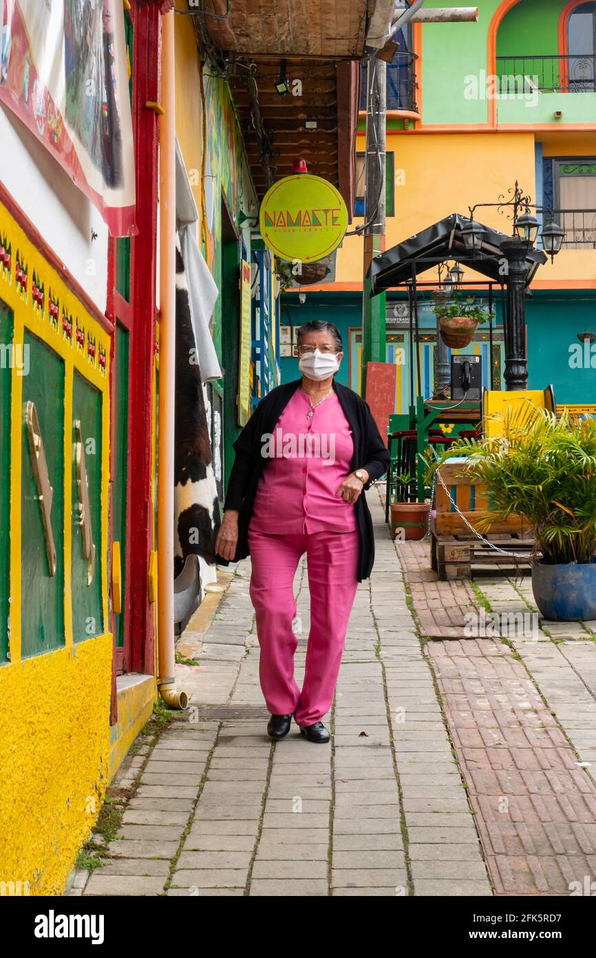 Guatapé, Antioquia, Kolumbien - 4 2021. April: Die alte Brünette, die mit Maske in Rosa gekleidet ist, geht den Straßenhang hinunter Stockfoto