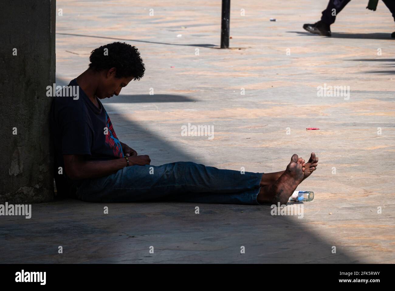 Medellin, Antioquia, Kolumbien - Januar 6 2021: Hispanischer Obdachloser sitzt an einer Betonwand und schaut traurig mit einer Alkoholflasche zu seinen Füßen herunter Stockfoto