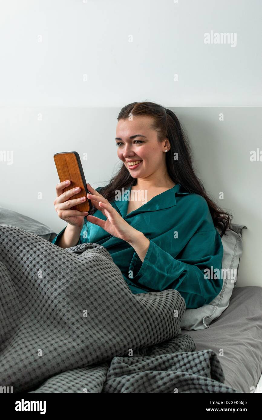 Der Seidenschlafanzug der jungen Frau, der im Bett liegt, hält ihr Handy an den Händen. Arbeiten Sie bequem von zu Hause aus. Wellness-Konzept. Stockfoto