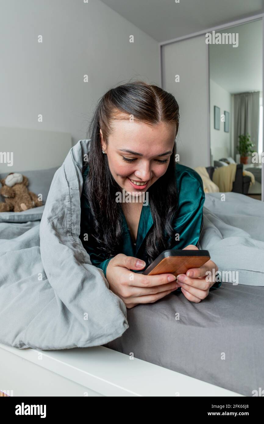 Der Seidenschlafanzug der jungen Frau, der im Bett liegt, hält ihr Handy an den Händen. Arbeiten Sie bequem von zu Hause aus. Wellness-Konzept. Stockfoto
