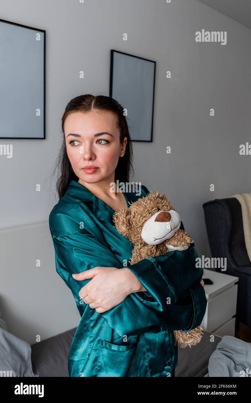 Eine junge Frau im Seidenpyjama sitzt auf dem Bett und umarmt einen Teddybären. Zurück zu den Wurzeln. Wellness-Konzept. Stockfoto